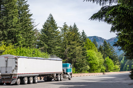 Old style cab over big rig industrial semi truck tractor transporting dusty cargo in covered low profile bulk semi trailer running on curving highway road with green mountain forest in Columbia Gorgeの写真素材