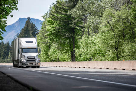 White big rig long haul industrial semi truck tractor transporting cargo in dry van semi trailer running on curving highway road with protective fence and green mountain forest in Columbia Gorgeの写真素材
