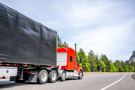 Red big rig long haul industrial semi truck tractor transporting cargo in dry van semi trailer running on curving highway road with protective fence and green mountain forest in Columbia Gorgeの写真素材