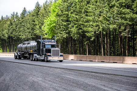 Black classic big rig industrial semi truck tractor transporting liquid cargo in stainless steel tank semi trailer running on curving highway road with green mountain forest in Columbia Gorgeの写真素材