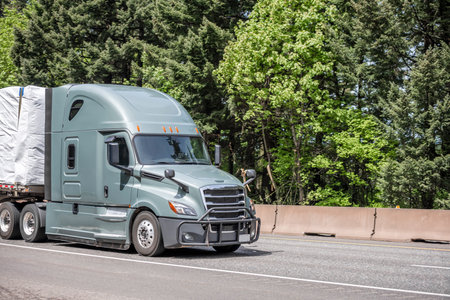 Gray long haul big rig industrial semi truck tractor transporting commercial cargo on flat bed semi trailer running on curving highway road with green mountain forest in Columbia Gorgeの写真素材