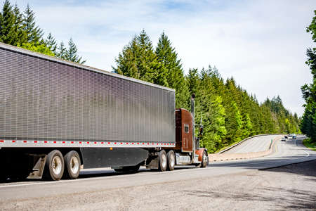 Brown big rig semi truck tractor with vertical chrome pipes transporting cargo in polished corrugated aluminum refrigerator semi trailer running on the green forest highway road in Columbia Gorgeの写真素材