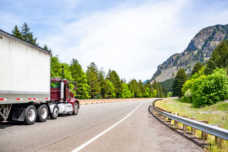 Burgundy day cab big rig industrial semi truck tractor for local deliveries transporting cargo in dry van semi trailer running on highway road with green forest trees in Columbia Gorgeの写真素材