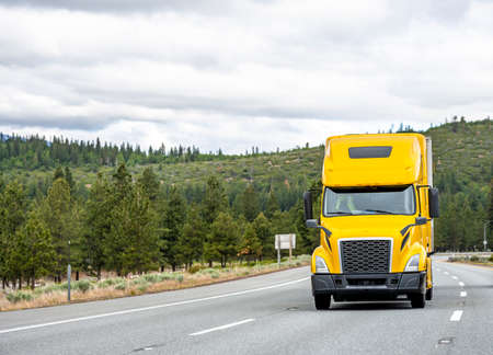 Popular Yellow bonnet big rig long haul industrial semi truck tractor transporting commercial cargo in dry van semi trailer running uphill on the winding mountain road with green forestの写真素材