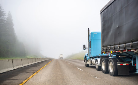 Blue big rig long haul semi truck transporting commercial cargo in dry van semi trailer running on turning one way highway road with forest on the hill at strong fog weather with poor visibilityの写真素材
