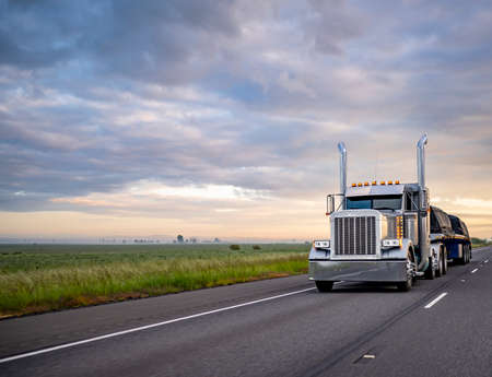Gray classic big rig American semi truck with turned on lights and chrome exhaust pipes transporting covered cargo on flat bed semi trailer driving on the twilight highway road with stormy clouds skyの写真素材