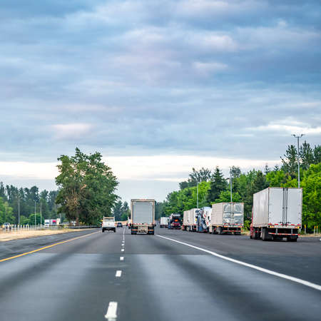 Group of big rigs semi trucks tractors transporting cargo in different semi trailers standing in a line by the side of the highway road for a break outside of the filled rest area parking lotの写真素材