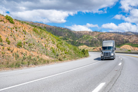 Classic gray bonnet powerful big rig long haul industrial semi truck tractor transporting commercial cargo in dry van semi trailer running uphill on the winding mountain road with rocks and forestの写真素材