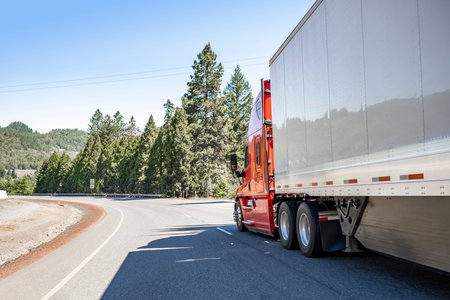 Red big rig long haul industrial semi truck tractor transporting commercial cargo in dry van semi trailer running for delivery on the summer Oregon mountain road with green trees on the hillsの写真素材