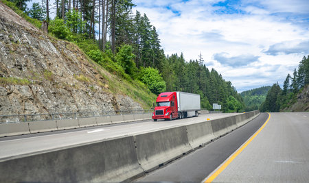 Red big rig long haul industrial semi truck tractor transporting commercial cargo in dry van semi trailer running for delivery on the summer Oregon mountain road with green trees on the hillsの写真素材