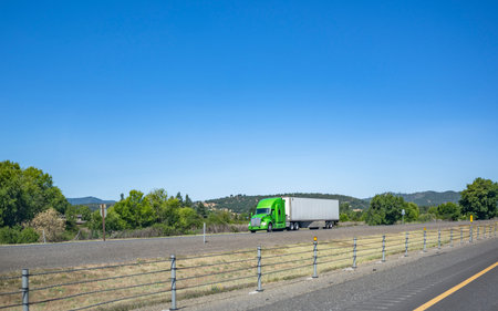 Green big rig long haul industrial semi truck tractor transporting commercial cargo in dry van semi trailer running for delivery on the summer highway road with rope fence in Californiaの写真素材
