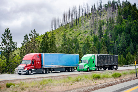Team of two big rigs industrial professional semi trucks tractors transporting cargo in dry van semi trailers driving side by side on the turning divided highway road along the forested mountainの写真素材