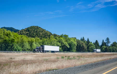 Team of two gray big rig semi trucks transporting commercial cargo in refrigerated semi trailers running in convoy on the one way highway road with grass dividing line and forest on the hillsの写真素材