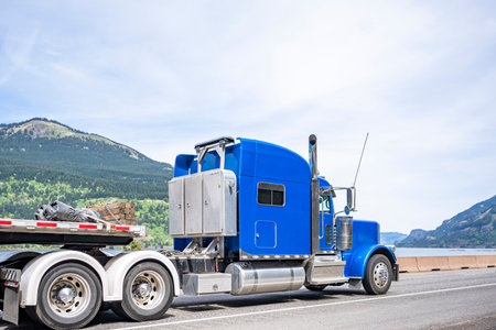 Industrial long hauler big rig blue semi truck tractor carrier transporting commercial cargo on step down semi trailer running on the winding highway road along Columbia river in Columbia Gorgeの写真素材