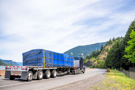 Industrial long hauler big rig blue semi truck tractor carrier transporting commercial cargo on flat bed semi trailer running on the winding highway road along Columbia river in Columbia Gorgeの写真素材