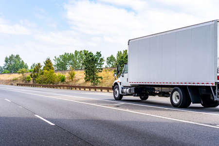 Classic medium duty day cab bonnet big rig white semi truck tractor with long box trailer for local deliveries transporting commercial cargo driving on the wide highway road with trees on the hillの写真素材