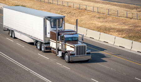 Industrial brown classic bonnet American idol big rig semi truck with chrome exhaust pipes transporting frozen commercial cargo in reefer semi trailer running on the summer divided highway roadの写真素材