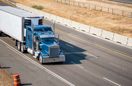Industrial blue classic bonnet American idol big rig semi truck with chrome exhaust pipes transporting frozen commercial cargo in reefer semi trailer running on the summer divided highway roadの写真素材