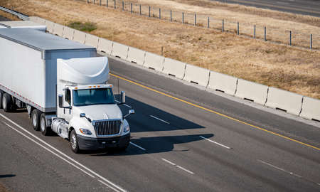 Day cab industrial bonnet white big rig semi truck with aerodynamic roof spoiler transporting commercial cargo in dry van semi trailer running on the summer highway road with yellow hillの写真素材