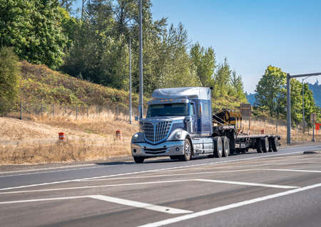 Stylish industrial blue bonnet professional big rig semi truck with truck driver rest compartment transporting commercial cargo on step down semi trailer running on the summer highway road entranceの写真素材