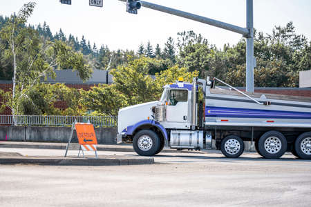 White stylish day cab big rig semi tip truck tractor transporting cargo in tipper semi trailer turning on the crossroad intersection with traffic lights and detour road signの写真素材