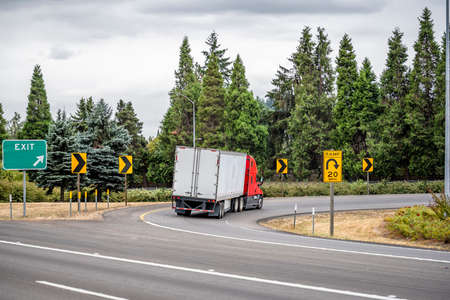 Industrial bonnet bright red classic big rig semi truck transporting commercial cargo in dry van semi trailer turning on the round highway exit with green trees on the side and road signsの写真素材