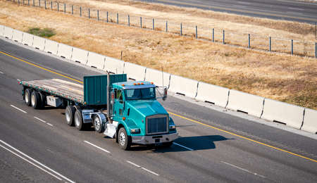 Classic old style bonnet day cab green semi truck for local freights transporting empty flat bed semi trailer with front protection wall running on the highway road to warehouse for the next loadの写真素材