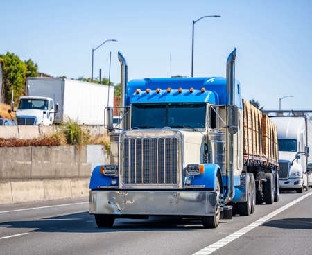 Classic heavy-duty industrial Blue Big rig classic semi truck with chrome accents transporting fastened lumber on flat bed semi trailer running on the highway road with another trucks traffic aroundの写真素材