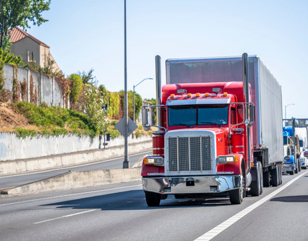 Red classic industrial big rig semi truck tractor transporting cargo in dry van semi trailer running on the divided highway road in front of another semi trucks going in the convoy lineの写真素材