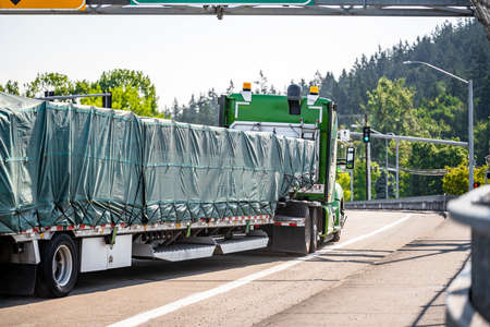 Green stylish bonnet big rig semi truck tractor with sleeping cab compartment for truck driver rest transporting cargo on the step down semi trailer turning on the city crossroad with traffic lightsの写真素材