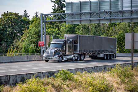 Industrial bonnet low cab black classic big rig semi truck with grille guard transporting commercial cargo in black dry van semi trailer running on the highway road exit with wrong way signの写真素材