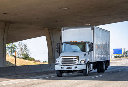 Industrial bonnet professional medium rig semi truck with chrome grill transporting commercial cargo in long box trailer running on the highway road under the concrete bridge across the roadの写真素材