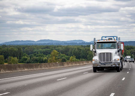Day cab bonnet white semi truck with additional roof lights and chrome exhaust pipes transporting cargo on flat bed semi trailer running on the one way highway road to warehouse for timely deliveryの写真素材