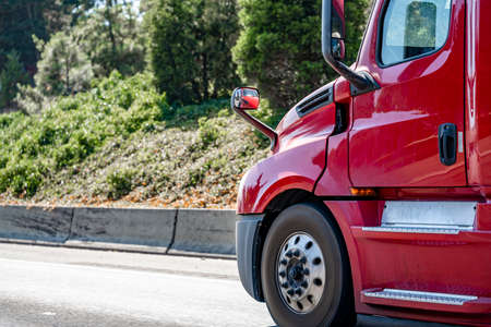Front part of the shiny bright red long hauler big rig semi truck tractor with high cab and aluminum accessories running on the road with forest on the hillsideの写真素材