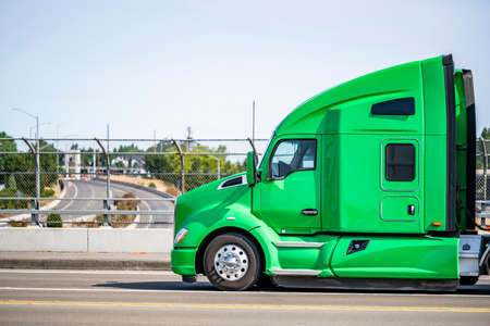 Green stylish bonnet big rig semi truck tractor with sleeping cab compartment for truck driver rest transporting cargo in black covered semi trailer running on the city street bridgeの写真素材