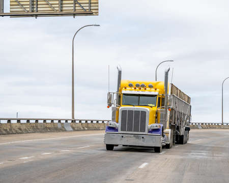 Big rig yellow semi truck with high cab transporting commercial cargo in open top bulk semi trailer running for delivery on the turning wide overpass highway roadの写真素材