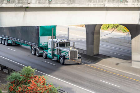Stylish classic American idol white and green semi truck tractor with high chrome pipes transporting cargo in covered semi trailer with front wall spoiler running on the highway road under the bridgeの写真素材