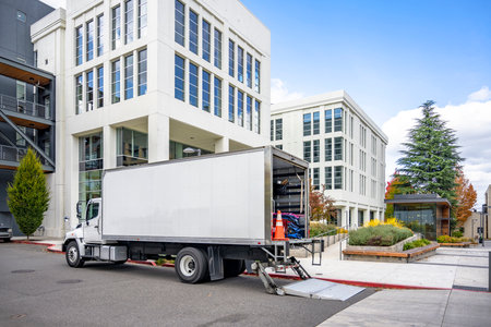 White middle class relocation rig day cab semi truck tractor with long spacious box trailer standing on the urban city street with multilevel apartment and office buildings unloading delivered goodsの写真素材