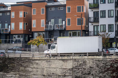 Compact Industrial professional white powerful middle class rig day cab semi truck tractor with long box trailer running for delivery on the urban city road with apartment and office buildingsの写真素材