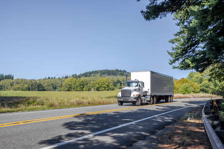 Classic big rig white bonnet semi truck with day cab transporting commercial cargo in dry van semi trailer running for delivery on the winding narrow road with forest and field on the sidesの写真素材