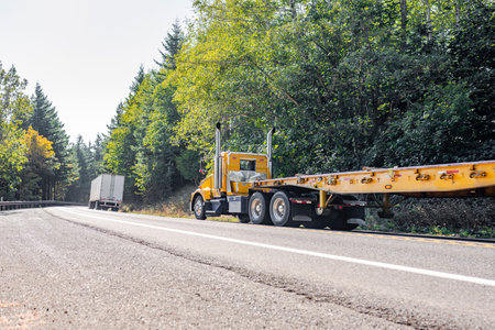 Convoy of two different big rig semi trucks with flat bed and dry van semi trailers running on the autumn highway road with forest on the sides to warehouse for the next freight loadの写真素材