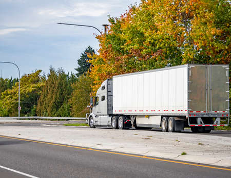 Classic big rig bonnet semi truck with grille guard transporting commercial cargo in dry van semi trailer running for delivery on the highway road with autumn yellow trees on the sideの写真素材