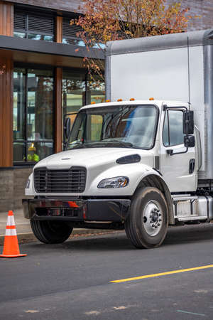 White middle class relocation rig day cab semi truck tractor with long spacious box trailer standing on the urban city street with multilevel apartment and office buildings unloading delivered goodsの写真素材