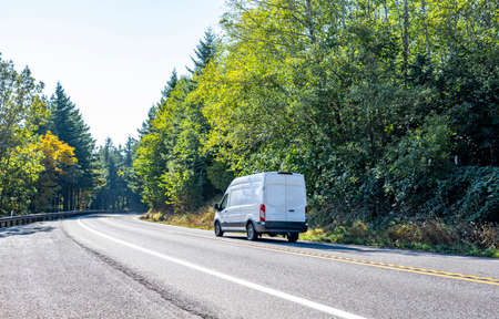 Compact small size white mini van with cargo compartment for local deliveries and small business needs for customers commercial cargo delivery driving on the turning road with forest on the sidesの写真素材