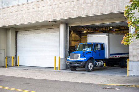 Blue middle class relocation rig day cab semi truck tractor with long spacious box trailer standing in dock inside of the multilevel apartment building in urban city unloading delivered goodsの写真素材