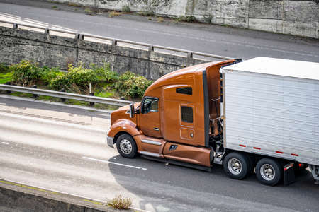 Classic big rig brown bonnet semi truck with high cab transporting commercial cargo in refrigerator semi trailer running for delivery on the multilevel highway intersectionの写真素材