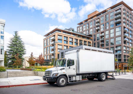 White middle class relocation rig day cab semi truck tractor with long spacious box trailer standing on the urban city street with multilevel apartment and office buildings unloading delivered goodsの写真素材