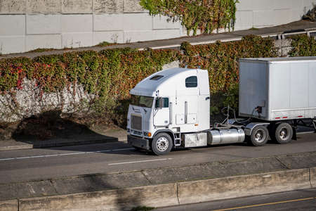 Old industry style over cab big rig white semi truck tractor transporting commercial cargo in dry van semi trailer moving on the divided highway road with upper entrance road and concrete wallの写真素材