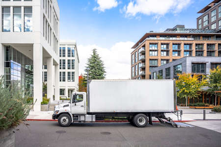 White middle class relocation rig day cab semi truck tractor with long spacious box trailer standing on the urban city street with multilevel apartment and office buildings unloading delivered goodsの写真素材