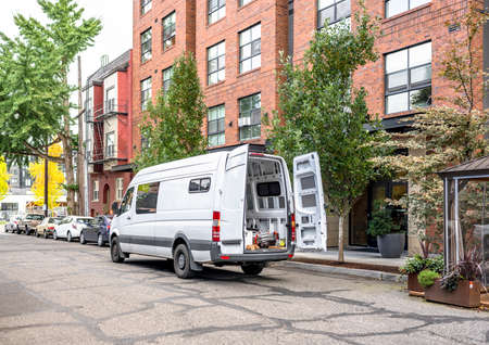 Commercial compact cargo small size white mini van delivered goods to client at multi-apartment high-rise building and standing on the autumn street of urban city waiting for unloadingの写真素材